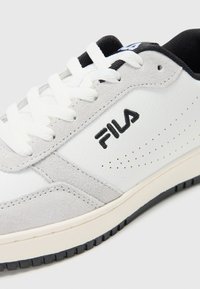 Hvite lærersneakers med gråsuede accenter, flate svarte snørebånd og en teksturert gummisåle med et svart mønster. Fila-logoen er vist.