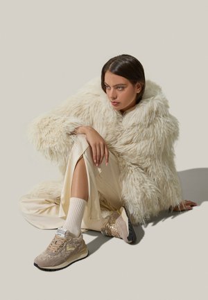 Sneakers beige con una toma strutturata e suola scura a contrasto, indossati con un cappotto in pelliccia finta color crema e pantaloni in satin chiaro. L'outfit include calze al ginocchio.