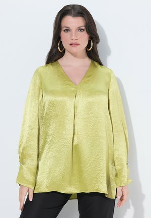 Nainen, jolla on pitkät tummat hiukset, suuri renkaankorvakorut ja kiiltävä limevihreä pitkähihaisen puseron sekä mustat housut valkoista taustaa vasten.