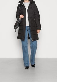 Abrigo acolchado negro con capucha, con botones a presión y cremalleras laterales, llevado sobre un suéter gris y jeans azules. Acentuado por un bolso estampado.
