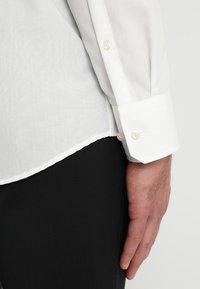 Camicia bianca a maniche lunghe realizzata in tessuto leggero, con colletto classico, bottoni perla e orlo curvo, abbinata a pantaloni neri.