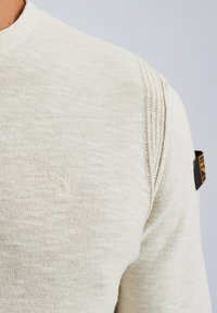 Svijetlosmeđi pleteni pulover s rebrastim ovratnikom i šavovima na ramenima. Sadrži diskretno izvezeni logo na prsima i tkanu oznaku na rukavu.