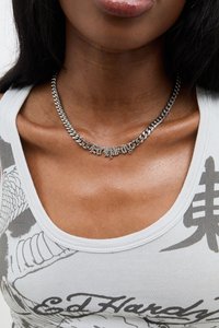 Colar de corrente prateada com um pendente decorativo que diz "Ed Hardy". Usado sobre uma camisola cinza claro com uma estampa gráfica.