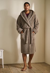 Peignoir en peluche gris avec col châle, poches latérales et ceinture à nouer. Texture douce avec une coupe jusqu'au genou. Porté par un modèle en intérieur.