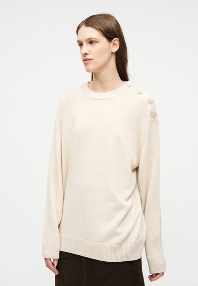 Γυναίκα με καστανά μαλλιά φοράει υπερμέγεθες κρεμ πουλόβερ με κουμπιά αστερίσκους στον αριστερό ώμο, συνδυασμένο με σκούρα φούστα, σε ουδέτερο φόντο.