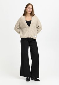 Young woman standing wearing beige buttoned cardigan, black wide-leg pants, black top, and black ankle boots on white background.