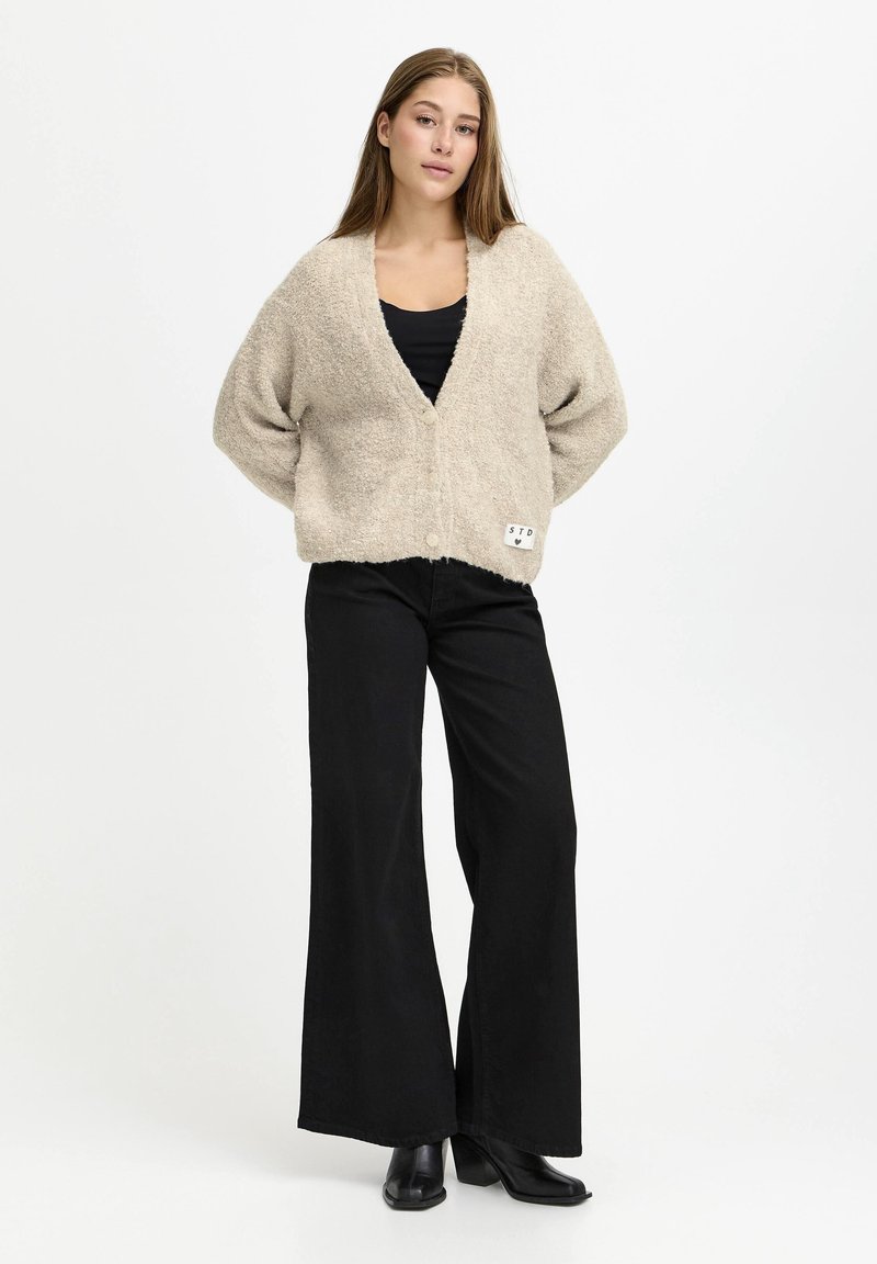 Young woman standing wearing beige buttoned cardigan, black wide-leg pants, black top, and black ankle boots on white background.