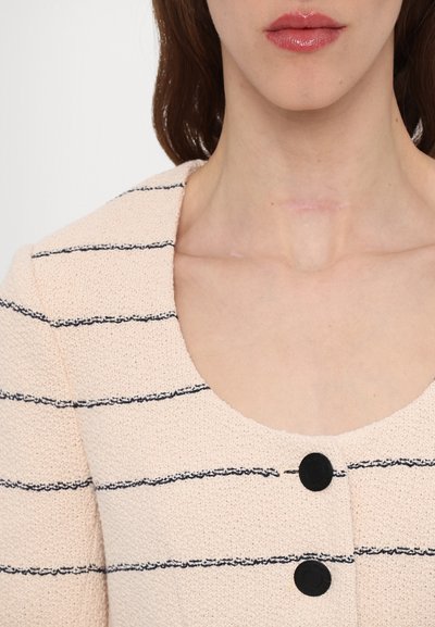 Lys beige cropped top med marineblå vandrette striber, rund halsudskæring og to sorte knapper på forsiden, der har et struktureret stof.