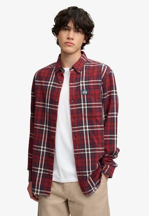 Junger Mann mit lockigem dunklem Haar, der ein rotes kariertes Hemd über einem weißen T-Shirt und beige Hosen trägt, steht mit einer Hand in der Tasche.