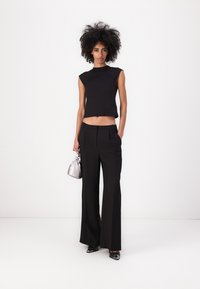 Top negro sin mangas con un cuello alto, combinado con pantalones anchos negros. La modelo sostiene un bolso plateado metálico y lleva zapatos negros.