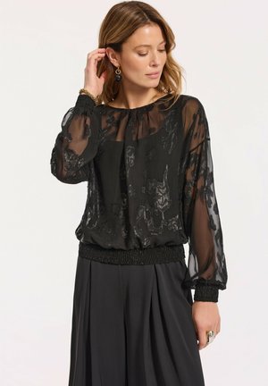 Kobieta dotykająca ucha, ubrana w czarną, przezroczystą bluzkę z długim rękawem w kwiatowy, błyszczący wzór oraz czarną spódnicę.