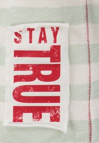 Testo rosso "STAY TRUE" su una toppa di tessuto bianco piegata, posizionata su un tessuto a righe verde chiaro e bianco con cuciture rosse lungo il bordo.