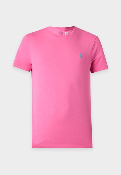 Απλό ροζ T-shirt με κοντά μανίκια και στρογγυλή λαιμόκοψη με μικρό μπλε κεντημένο λογότυπο στο αριστερό στήθος σε μονόχρωμο φόντο.