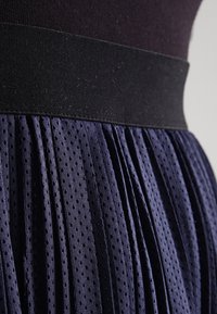 Marinblå plisserad kjol i en texturerad tyg med små hål, kompletterad med ett svart elastiskt midjeband för ökad komfort och passform.