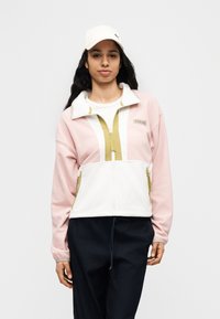 Γυναίκα με λευκό καπέλο, ροζ και λευκό ζακέτα φλις με φερμουάρ και σκούρο παντελόνι, στέκεται μπροστά σε απλό ανοιχτό φόντο.