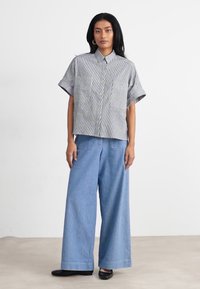 Camicia a maniche corte a righe in tessuto blu e bianco, abbinata a pantaloni in denim chiaro a gamba larga e scarpe nere. Il modello si trova su uno sfondo neutro.