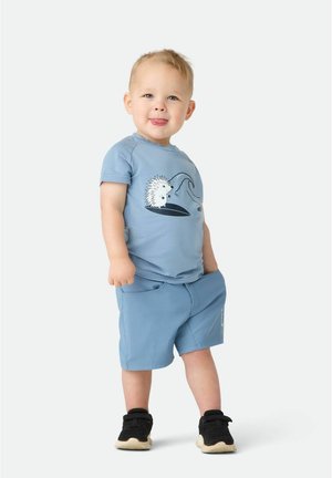 Småbarn i lyseblå trøje med pindsvineprint og matchende shorts står med hænderne i lommerne iført sorte sneakers.