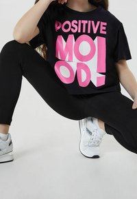Černé tričko s tučnými růžovými a bílými nápisy "POZITIVNÍ NÁLADA", spárované s černými legínami a bílými teniskami. Ležérní, uvolněný střih.