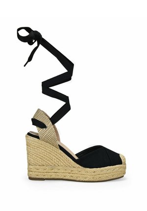 Sandalia cuña negra de lona con suela de yute, con una correa de tobillo de cinta negra trenzada y un acento en el tacón texturizado en beige.