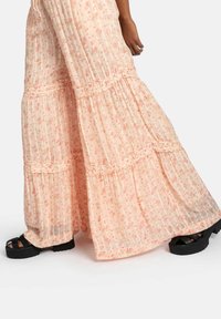 Robe maxi fleurie rose avec des volants superposés, fabriquée en tissu léger. Sandales à plateforme noires visibles en bas, avec un design à lanières.