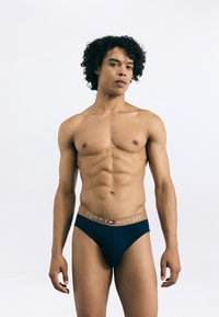 Modré bavlnené slipy so zlatým logom na páse s nápisom "TOMMY HILFIGER", ktoré ukazujú hladký, priliehavý dizajn. Model stojí pred jednofarebným pozadím.