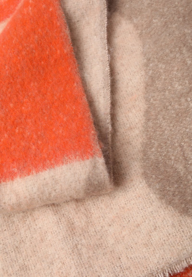 Miękki wełniany koc w wyrazistych odcieniach pomarańczowego i taupe, z teksturowaną powierzchnią i frędzlami, tworzący kontrastowe kolory i wzory.