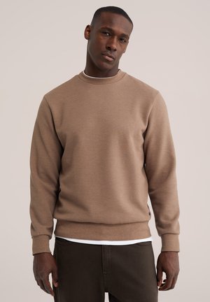 Homme portant un pull texturé marron clair à manches longues sur une chemise blanche et un pantalon marron foncé, debout devant un fond uni.