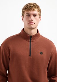 Bruine fleece pullover met een half-rits kraag, voorzien van een textuurstof en een zwart hexagonaal logo op de linkerkant van de borst.