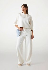 Weißer kurzer Hoodie mit einer Fronttasche, kombiniert mit weißen, weit geschnittenen Hosen. Weicher Stoff mit subtiler Textur. Hält eine hellblaue Kappe.