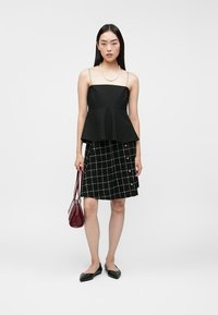 Czarny top peplum z cienkimi ramiączkami, zestawiony z czarną spódnicą sięgającą do kolan, z białym wzorem kratki i guzikami. Trzyma bordową torebkę.
