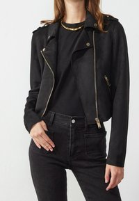 Giubbotto da biker in ecopelle nera con zip dorata, tasca laterale e spalline, abbinato a una maglia nera e jeans.