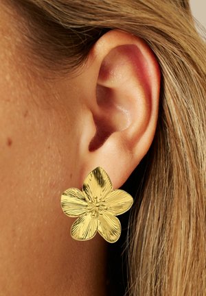 Goldfarbenes, blumenförmiges Ohrstecker mit strukturierten Blütenblättern und einem zentralen Blumendetail, getragen am Ohr, neben glatter Haut und Haaren.