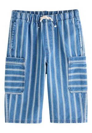 Jeune garçon souriant tenant un petit ours en peluche bleu, portant un t-shirt blanc, un pantalon bleu rayé et des baskets blanches.