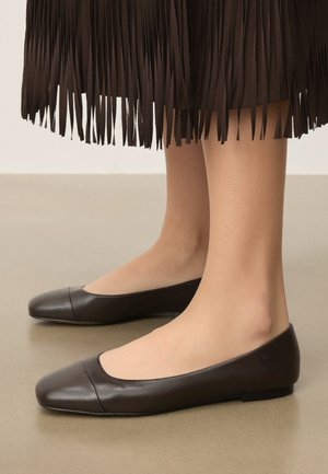 Brune læderballet flats båret med et mørkebrunt nederdel med frynser, sat mod en neutralfarvet gulv- og vægbaggrund.