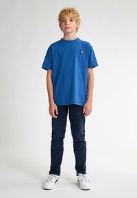 Camiseta de algodón azul con cuello redondo y mangas cortas, con un pequeño parche de logo en el pecho. Combinada con vaqueros oscuros y zapatillas blancas.