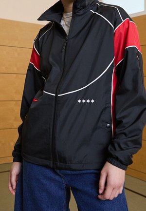 Person trägt eine schwarz-rote Reißverschlussjacke mit weißen Paspeln und vier weißen Sternen auf der Vorderseite, kombiniert mit blauen Jeans.