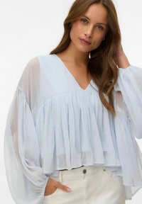 Femme aux longs cheveux bruns portant un chemisier plissé bleu clair transparent à manches volumineuses et un pantalon blanc, posant devant un fond blanc.