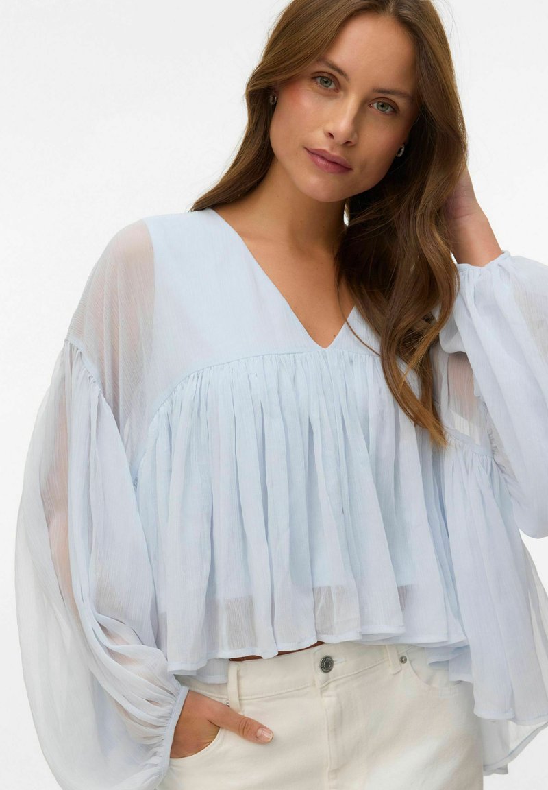 Femme aux longs cheveux bruns portant un chemisier plissé bleu clair transparent à manches volumineuses et un pantalon blanc, posant devant un fond blanc.