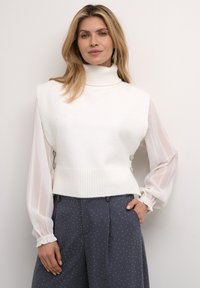 Biely sveter s vysokým golierom a bez rukávov s rebrovaným spodným okrajom, spárovaný s priesvitnou bielou blúzkou s dlhým rukávom. Šedé nohavice s bodkami dotvárajú vzhľad.