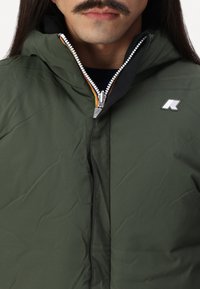 Mand med mørkegrøn hættejakke med synlig lynlås og lille hvid-grå logo på brystet, sort skjorte under.