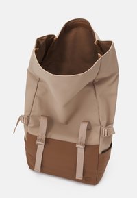 Sac à dos beige et marron avec ouverture sur le dessus, deux sangles avant à boucle et poches latérales, posé en position verticale sur un fond blanc.