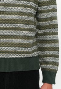 Zielony, biały i musztardowy sweter w paski z dzianiny, z ribbowymi mankietami i dolnym wykończeniem, noszony z czarnymi spodniami sztruksowymi, ścięty w talii i nad nadgarstkiem.