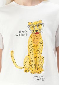Hvit t-skjorte med et gult leopardmotiv med svarte prikker, iført briller og røde lepper. Teksten sier "BAD VIBES" og "DON'T GO WITH MY LIPSTICK."