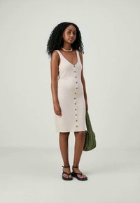 Mulher com cabelo encaracolado veste um vestido bege sem mangas com botões, sandálias pretas, um colar de conchas e segura uma bolsa verde texturizada.