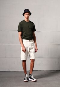 T-shirt vert à manches courtes, shorts blancs et chapeau seau noir, associés à des baskets montantes noires avec semelles blanches, le tout sur un fond neutre.