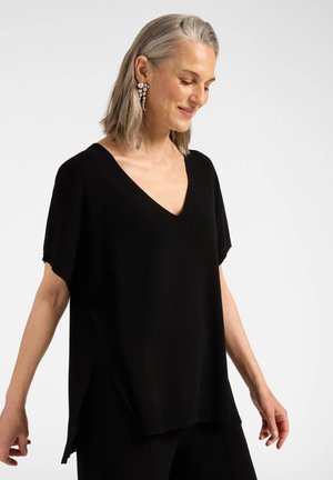 Femme d'âge moyen aux cheveux gris portant des boucles d'oreilles en perles et un t-shirt noir décontracté à col en V, souriante, regardant vers le bas et étendant légèrement les bras.