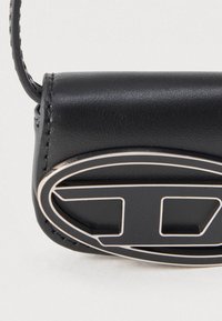 Juodas odinis krepšio kampas, siūtas dirželis ir ovalo formos juodos ir smėlio spalvos metalinis logotipo detalė.