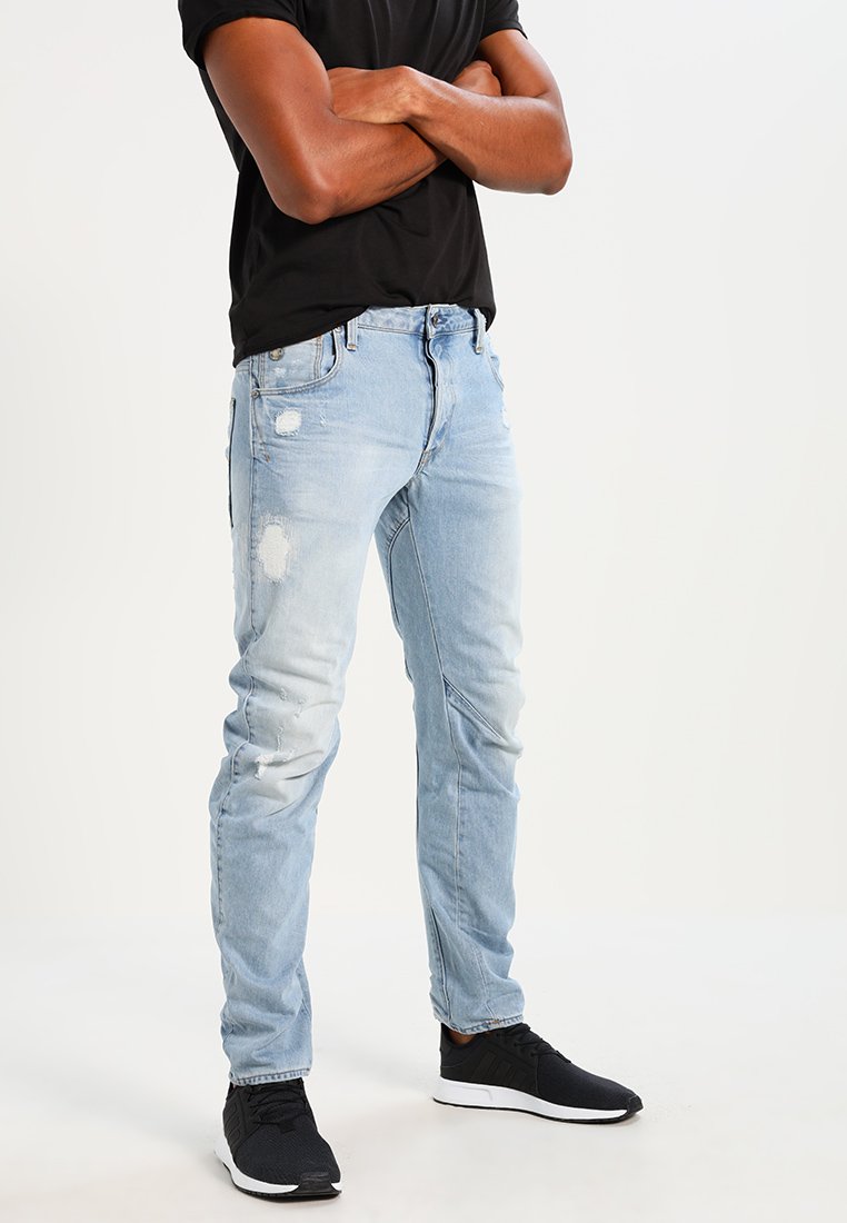 Jeans desgastados azul claro com um corte slim, apresentando rasgos nos joelhos, design de cinco bolsos e fecho de botão. Usados com ténis pretos.