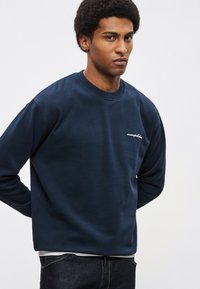Tmavomodrá mikina z mäkkej, textúrovanej látky s okrúhlym výstrihom a vyšívaným logom na hrudi. Trochu voľnejší strih.