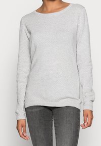 Pull gris clair en mailles texturées. Encolure ronde, manches longues et ourlet côtelé. Associé à un jean gris foncé.